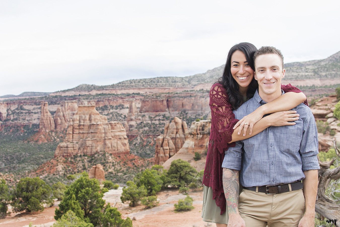 Engaged couple standing before a canyon with her arms around his shoulders