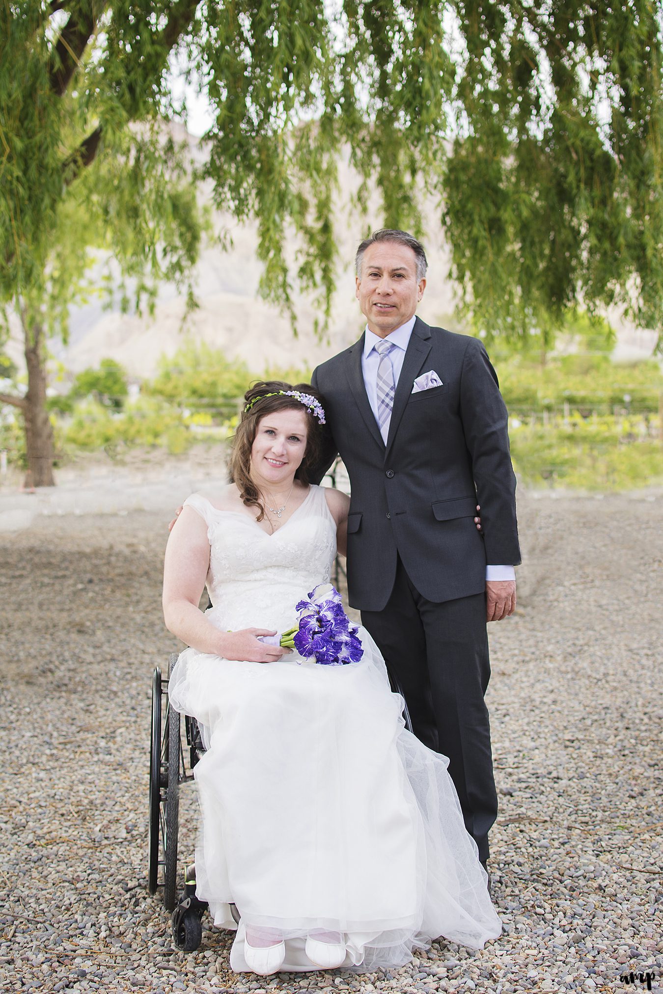 Bride in wheelchair and Groom standing, together beneath a willow tree