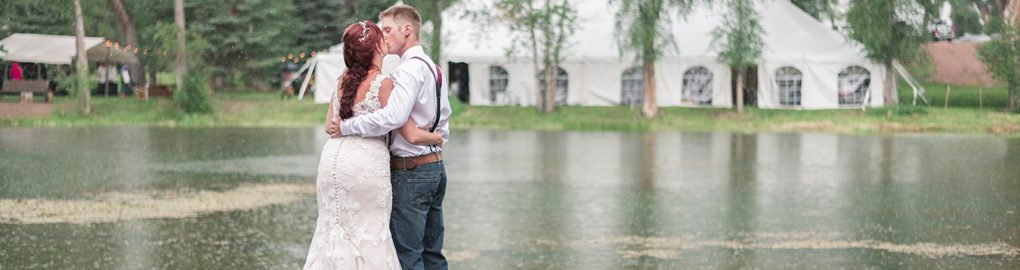 Bride and groom kissing on the dock of a pond with wedding in background