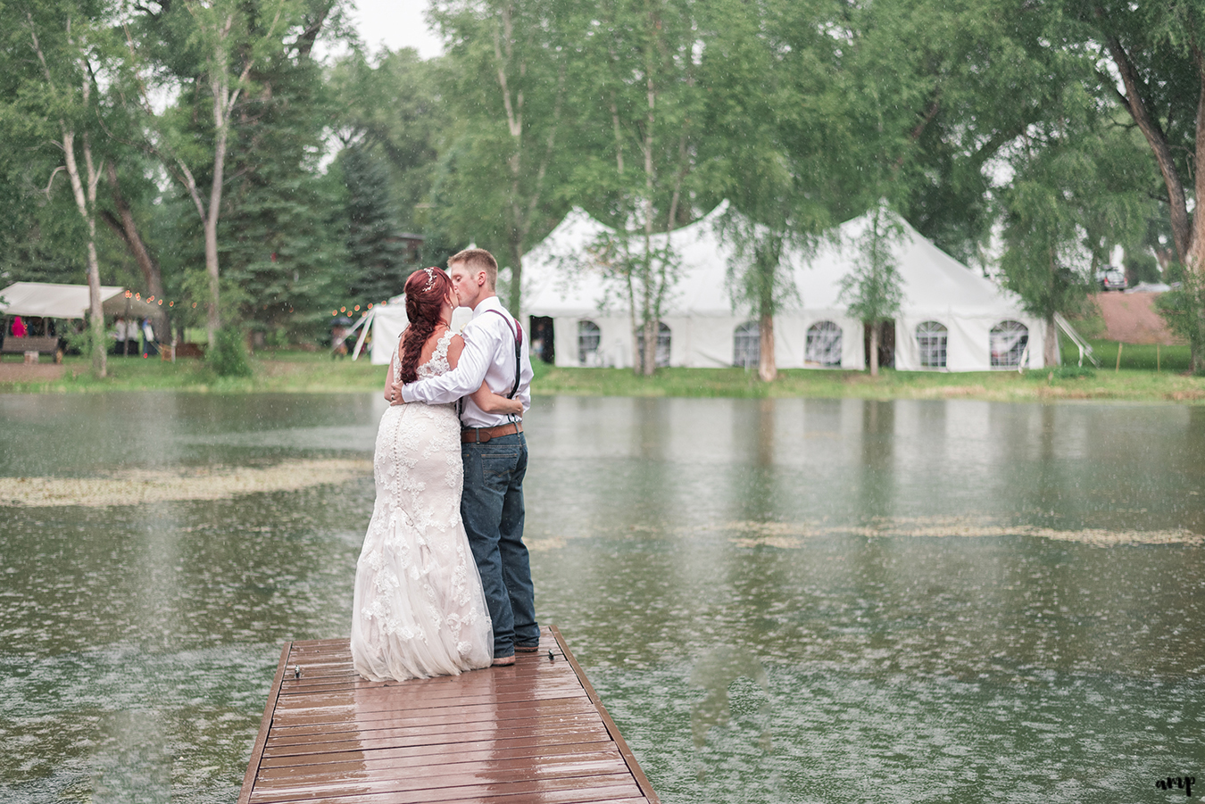 Bride and groom kissing on the dock of a pond with wedding in background