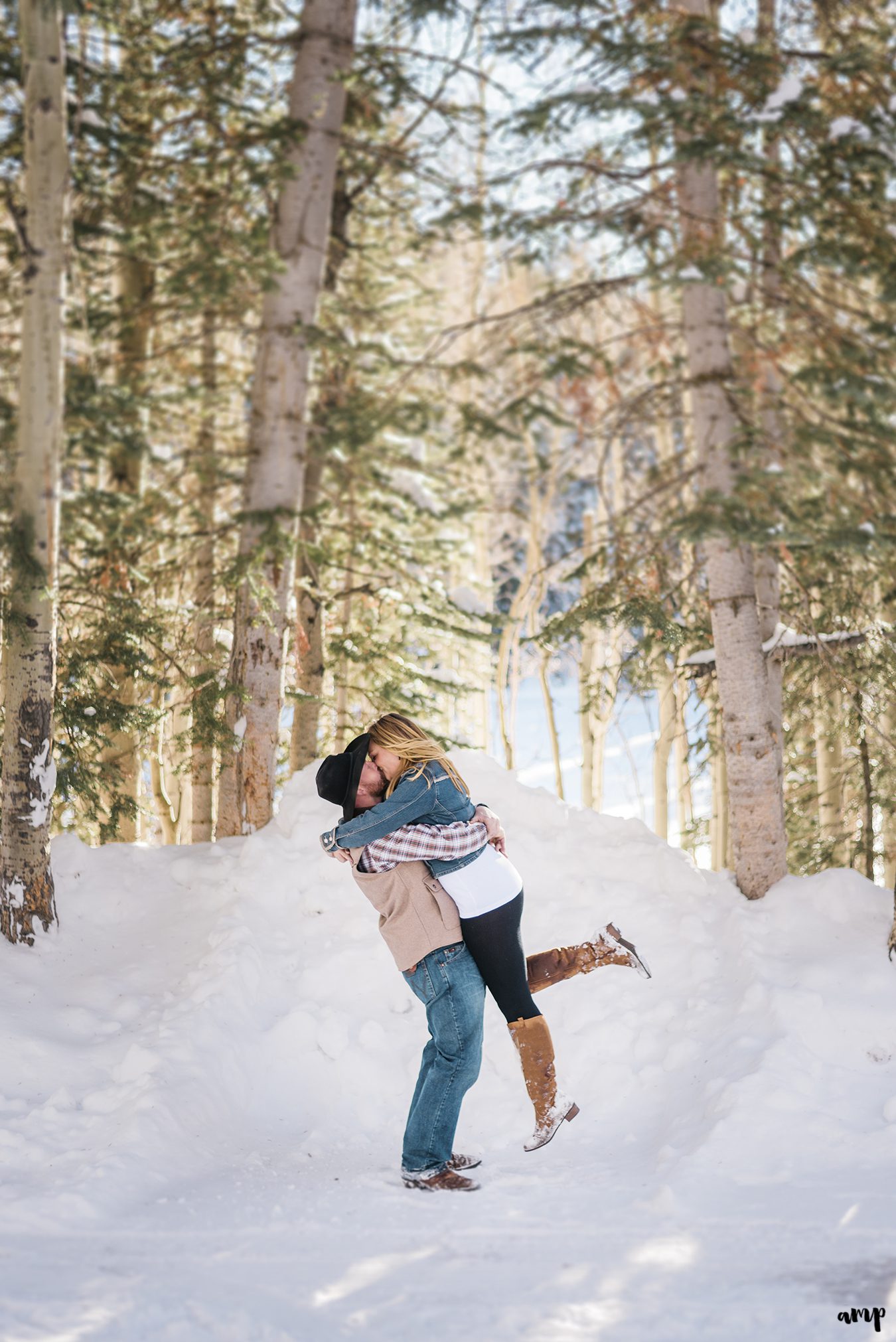 Engaged couple kissing beneath snowy pine
