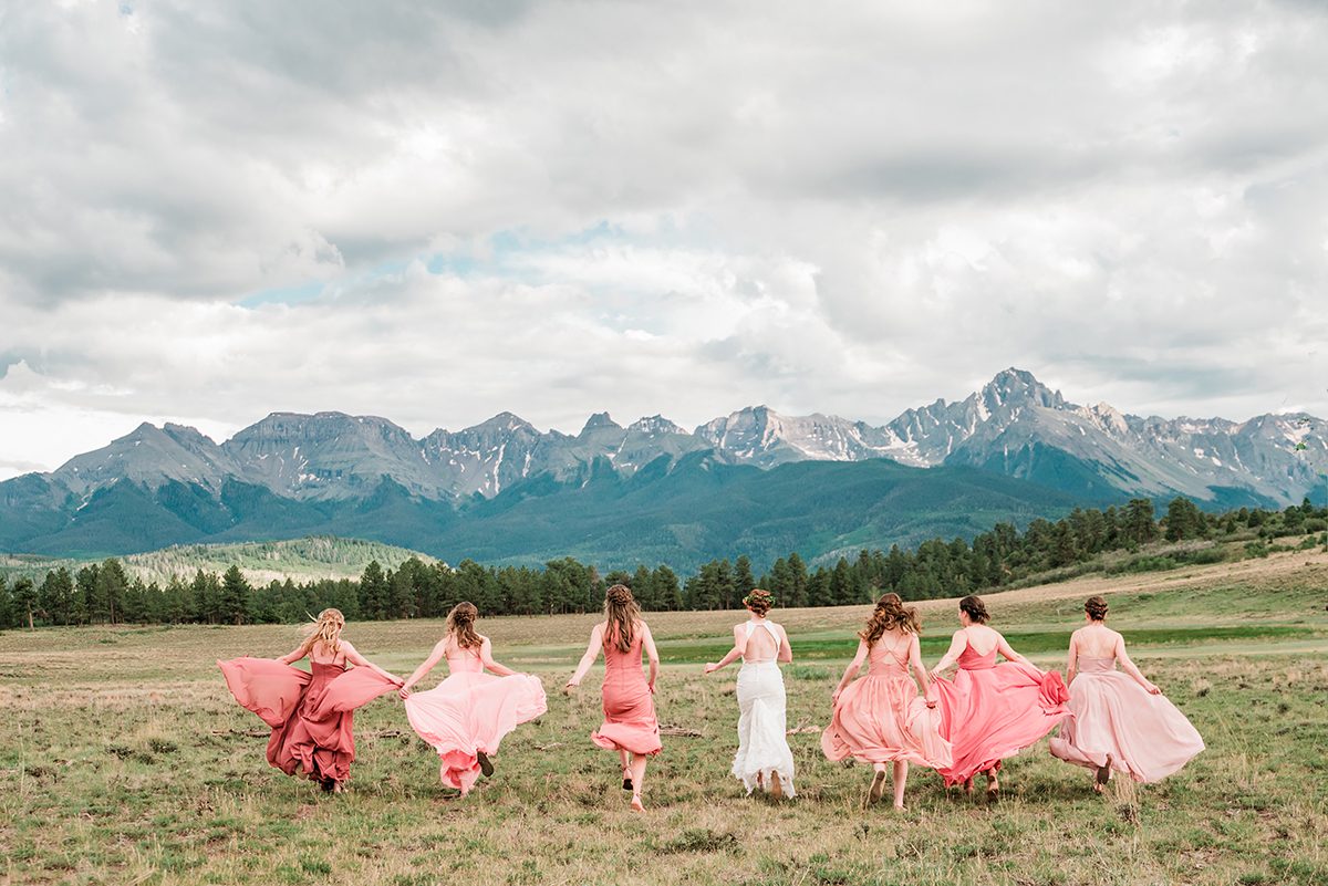 Bridesmaids and Sarah run through the meadow with their pink dresses flowing in the wind
