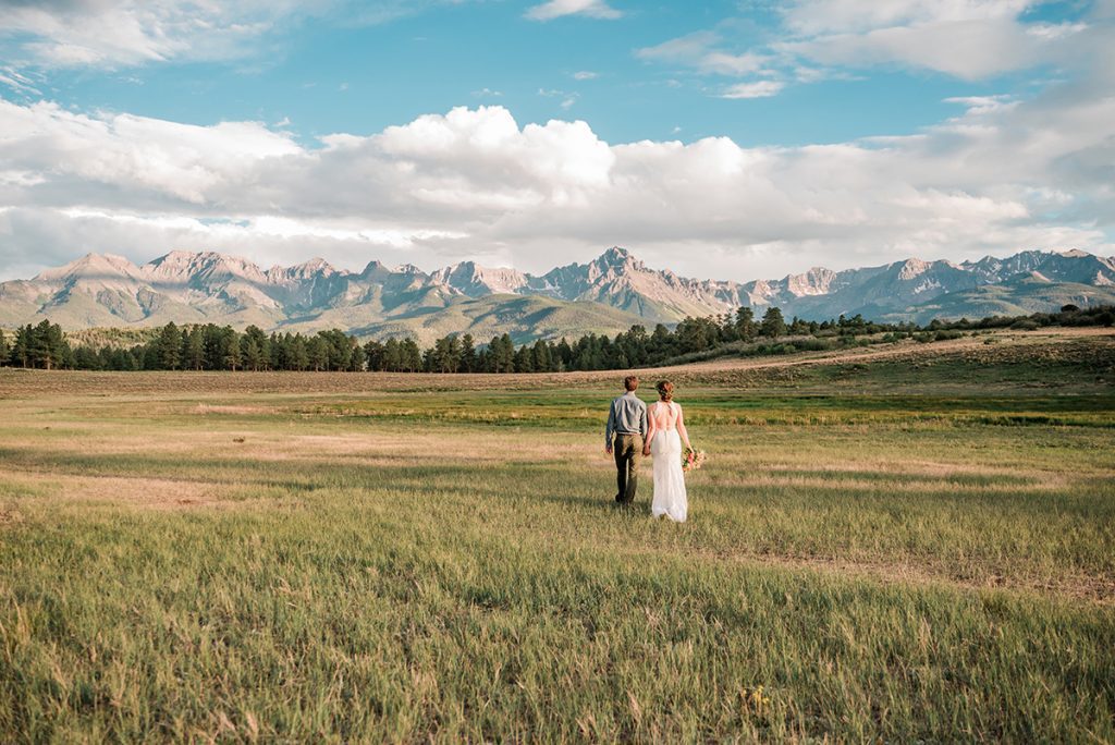 Sarah & Tom walk hand-in-hand through the meadow with the mountains along the horizon
